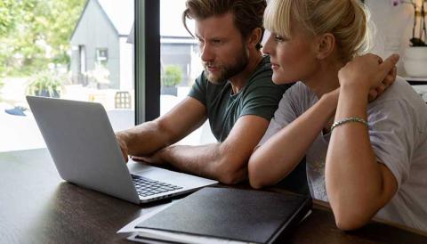A man and a woman sit at a table looking at a laptop together